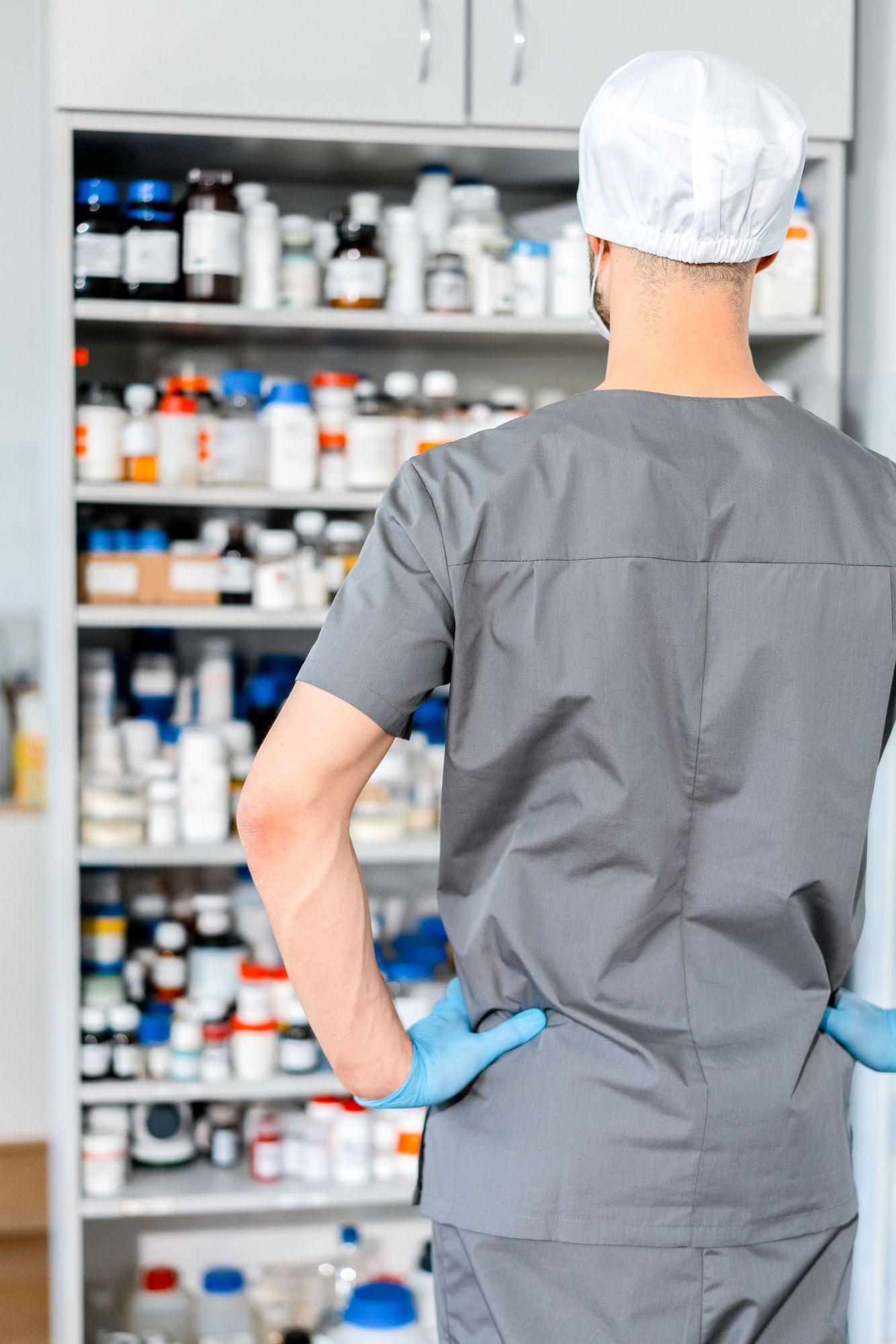 Préparateur en tenue grise et charlotte, vu de dos, devant une armoire de flacons de médicaments.
