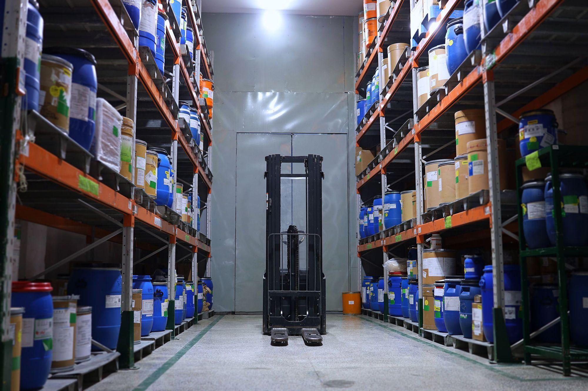 Allée d’un entrepôt pharmaceutique : rayonnages chargés de fûts et de cartons, chariot élévateur au fond.