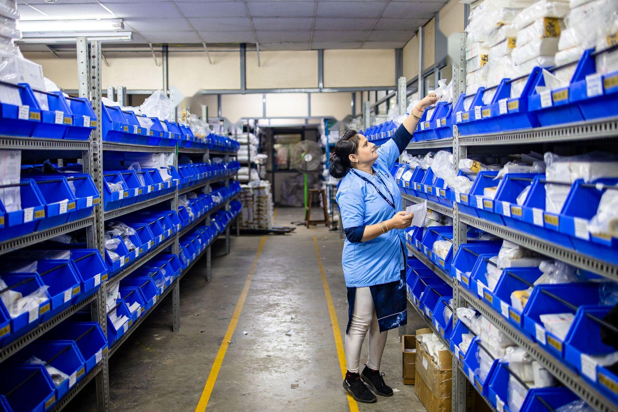 Préparatrice vérifiant une fiche de commande entre des rayonnages de bacs bleus.